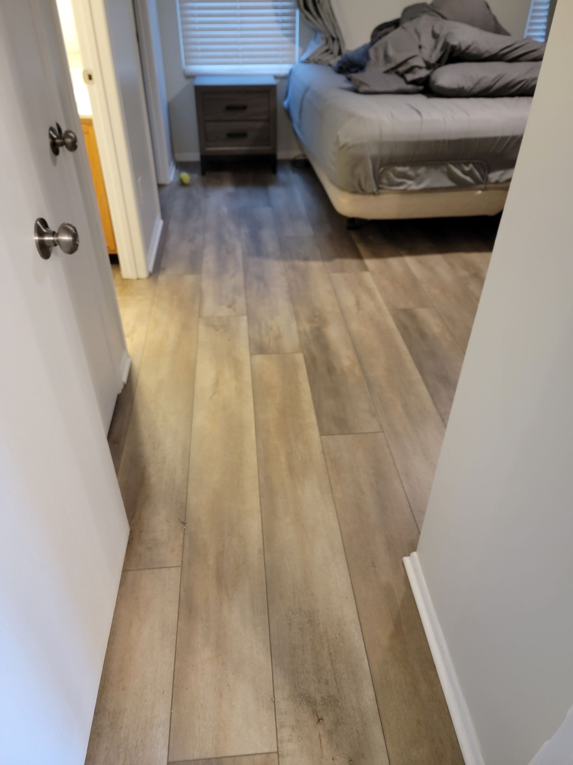 Narrow hallway with wood-look flooring leading to a bedroom with an unmade bed and nightstand.