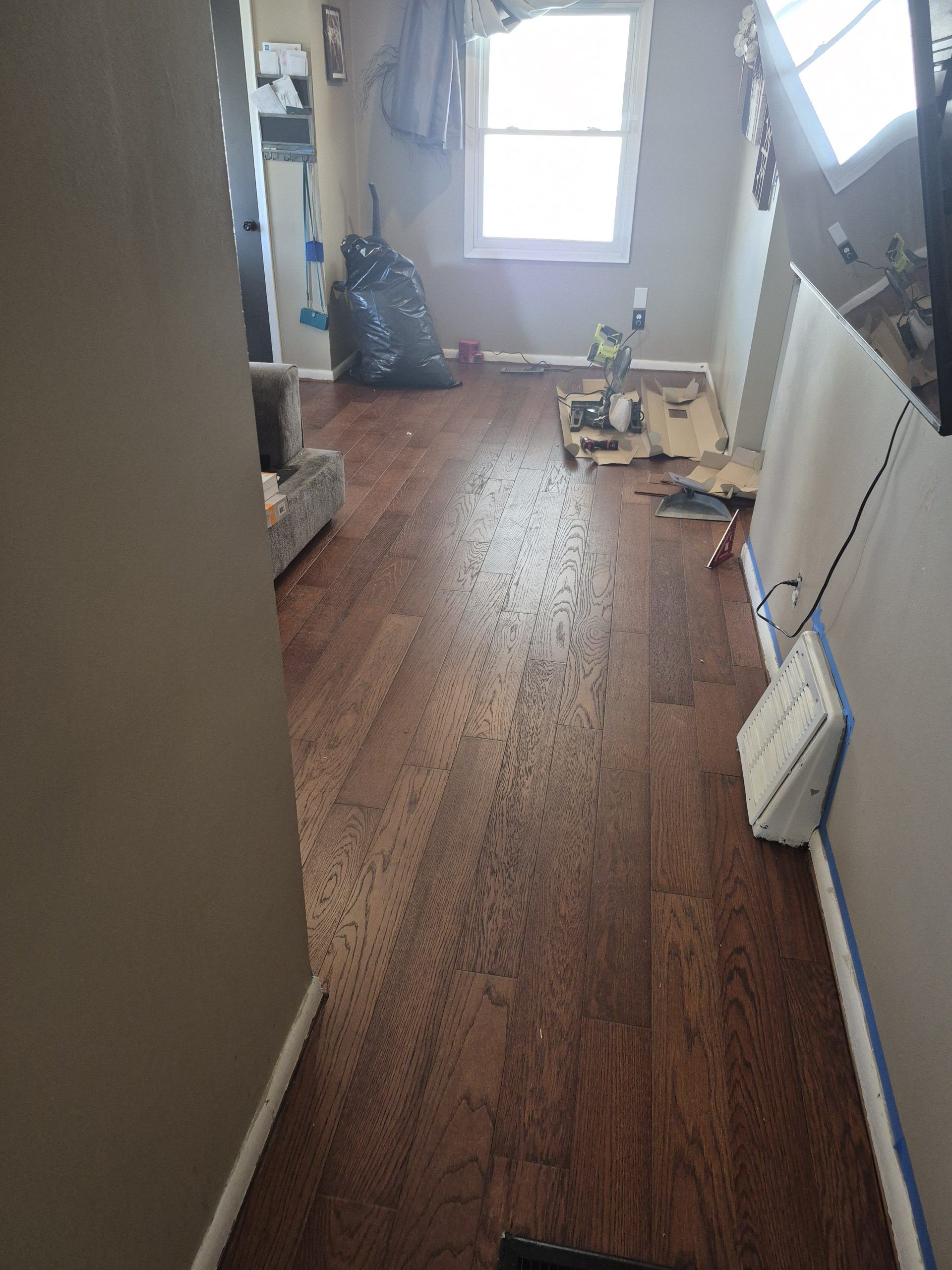 Narrow hallway with wood floor, white walls, a window, and tools and materials scattered along the right side.