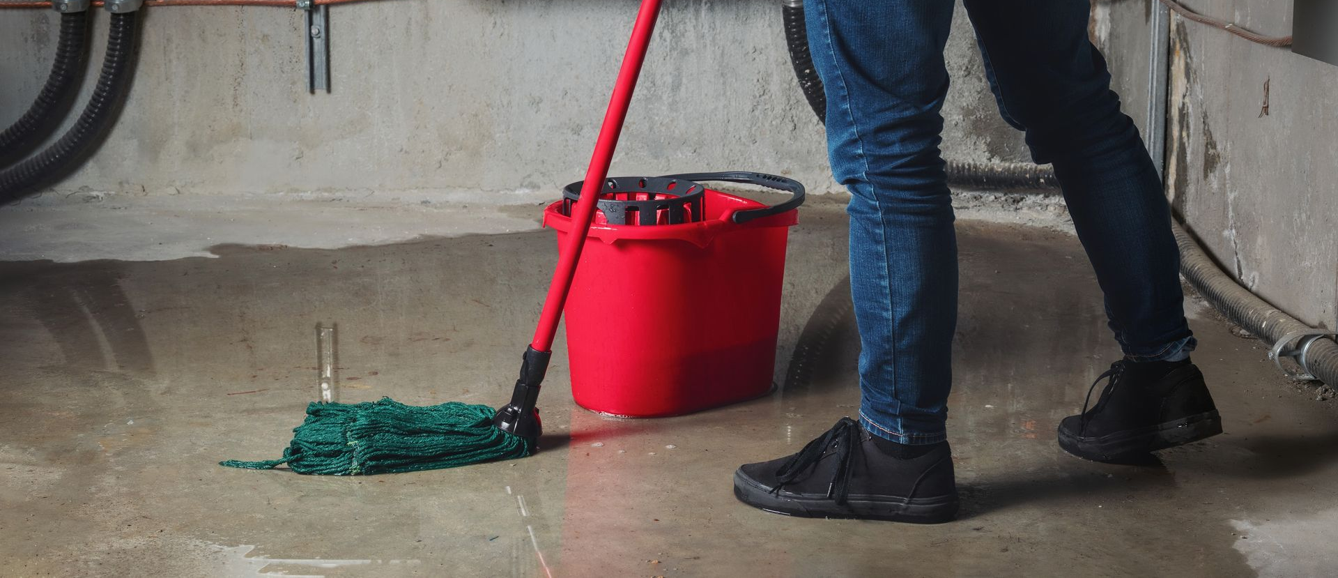 Person mopping a wet garage floor beside a red bucket and parked car tires