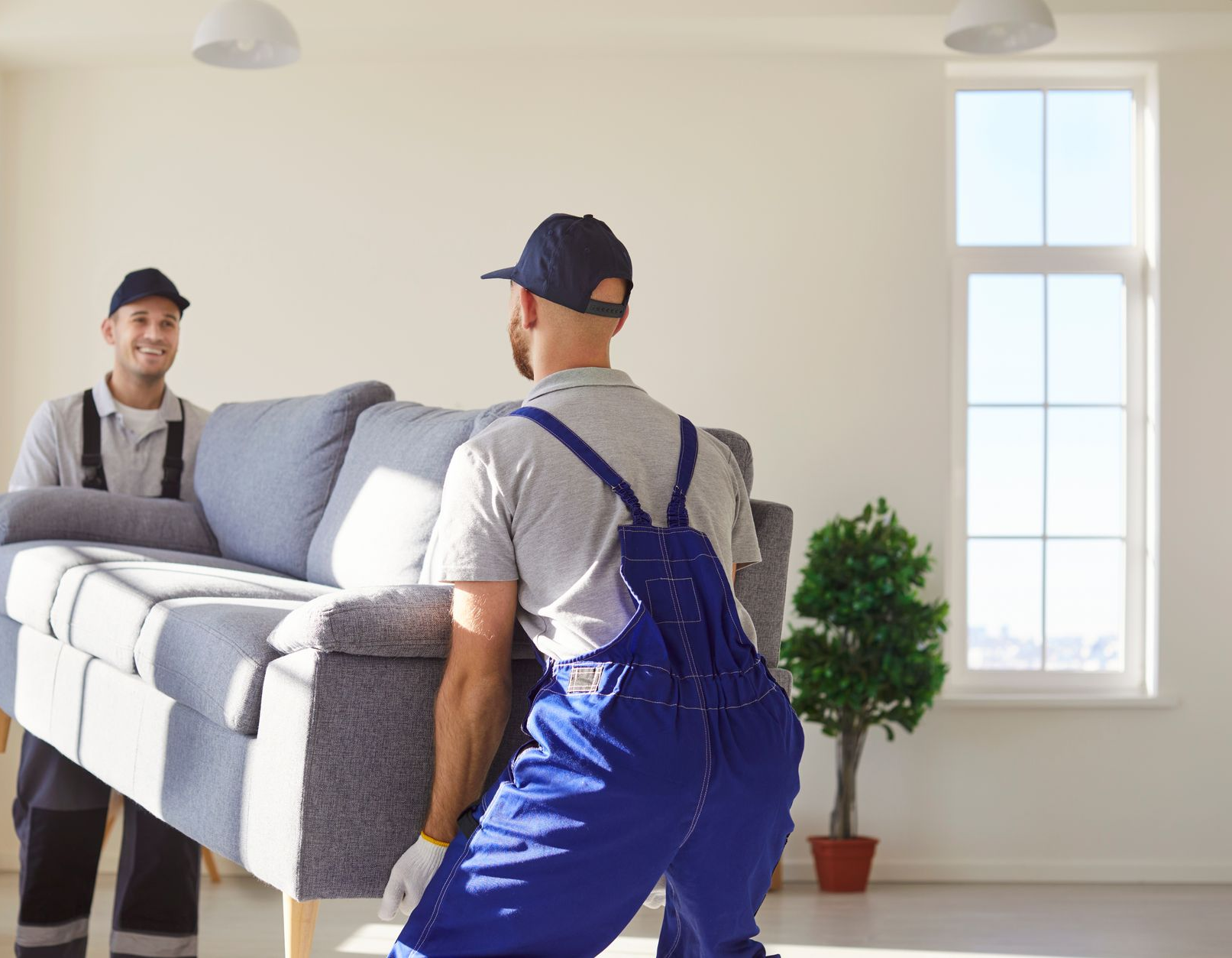 Two movers carrying a gray sofa through a bright room near a window