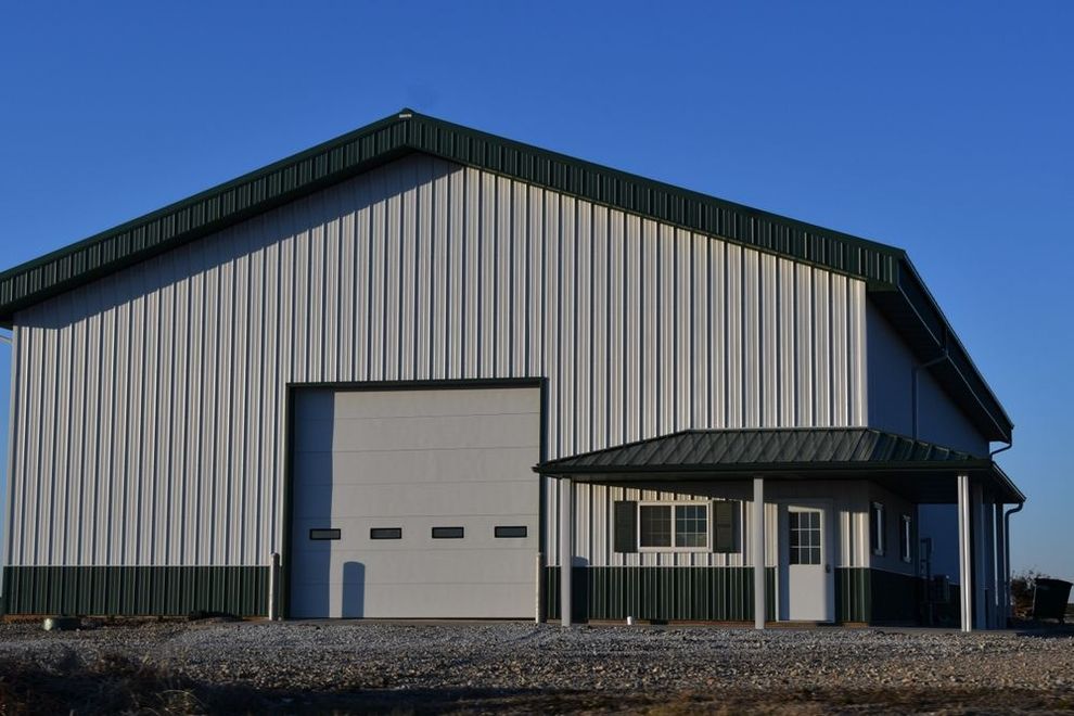 Large gray metal warehouse with a dark roof and small attached entrance building on a gravel lot