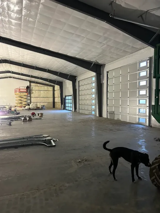 Inside a large empty industrial warehouse with a black dog near the foreground and stacked items in back