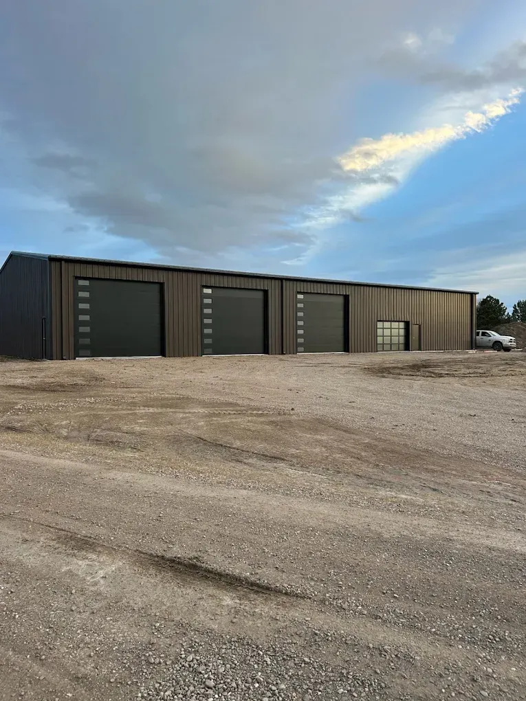 Wide black metal industrial building beside a gravel lot under a cloudy sky