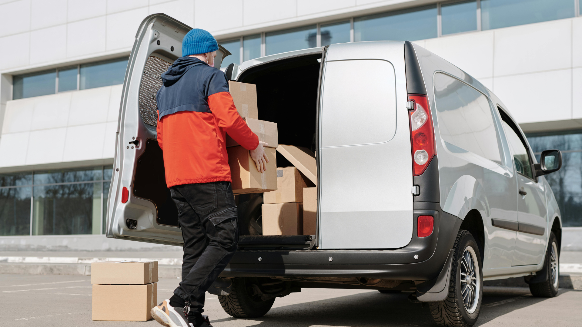 Delivery person loading cardboard boxes into a silver van with the rear door open, outdoors.