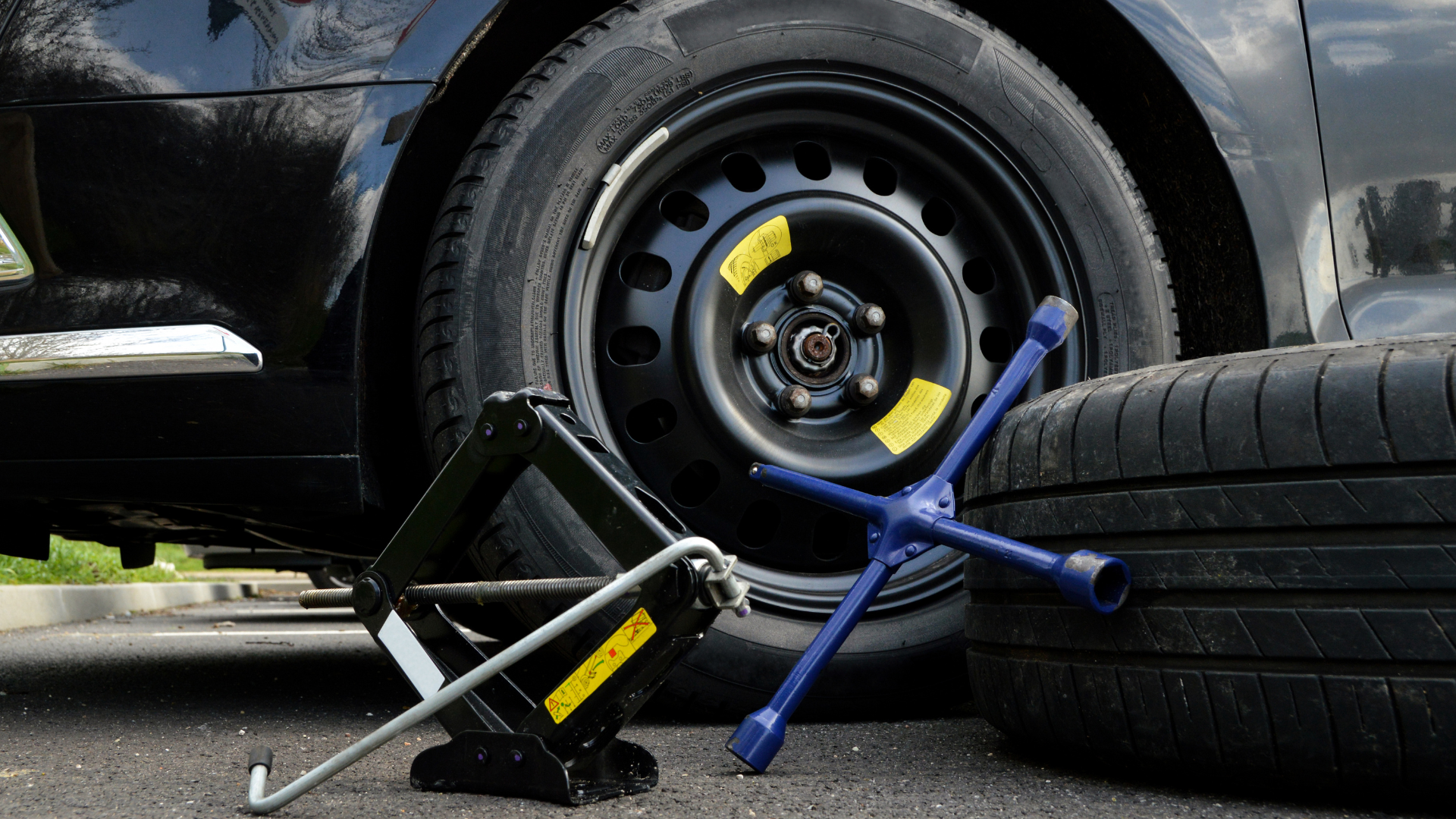 tools to fix a flat tire, leaning against a fresh tire with a car with a flat tire behind
