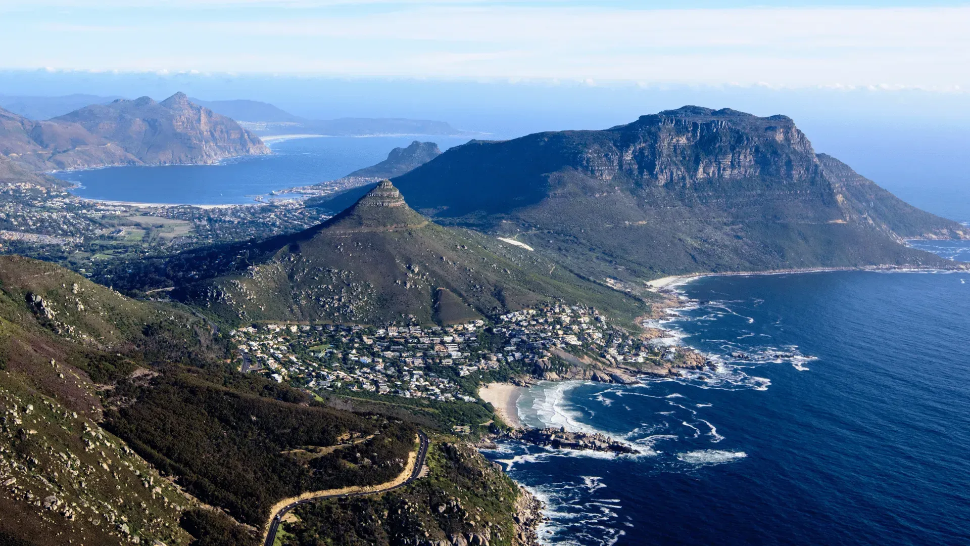 Coastal mountain overlooking a blue bay with a town and winding shoreline, seen under a clear sky