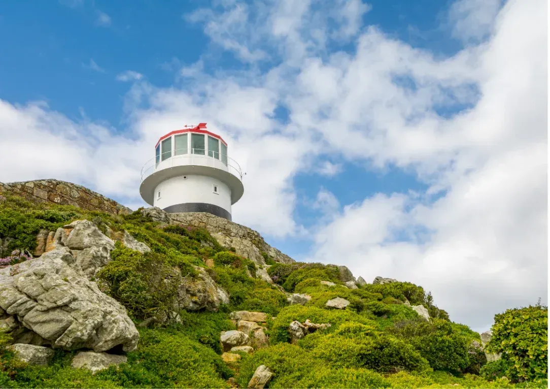 White lighthouse on a rocky green hillside under a blue sky with clouds