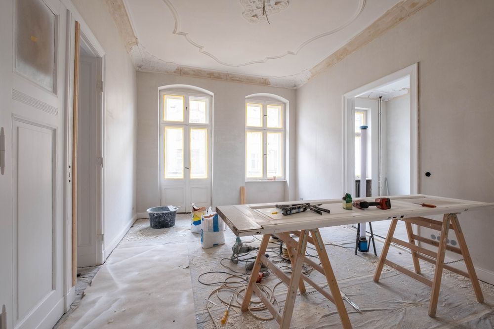 Interior room undergoing renovation, with open door, two windows, tools on sawhorses, and paint supplies.