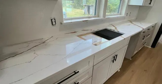 White kitchen countertop with gold veining, sink, and cabinets below a window.