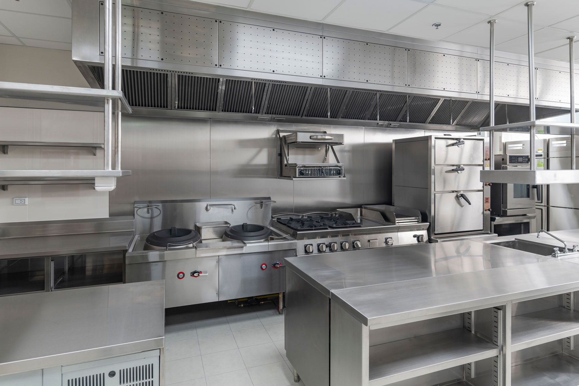Stainless-steel commercial kitchen with stoves, prep counters, shelves, and ventilation hood