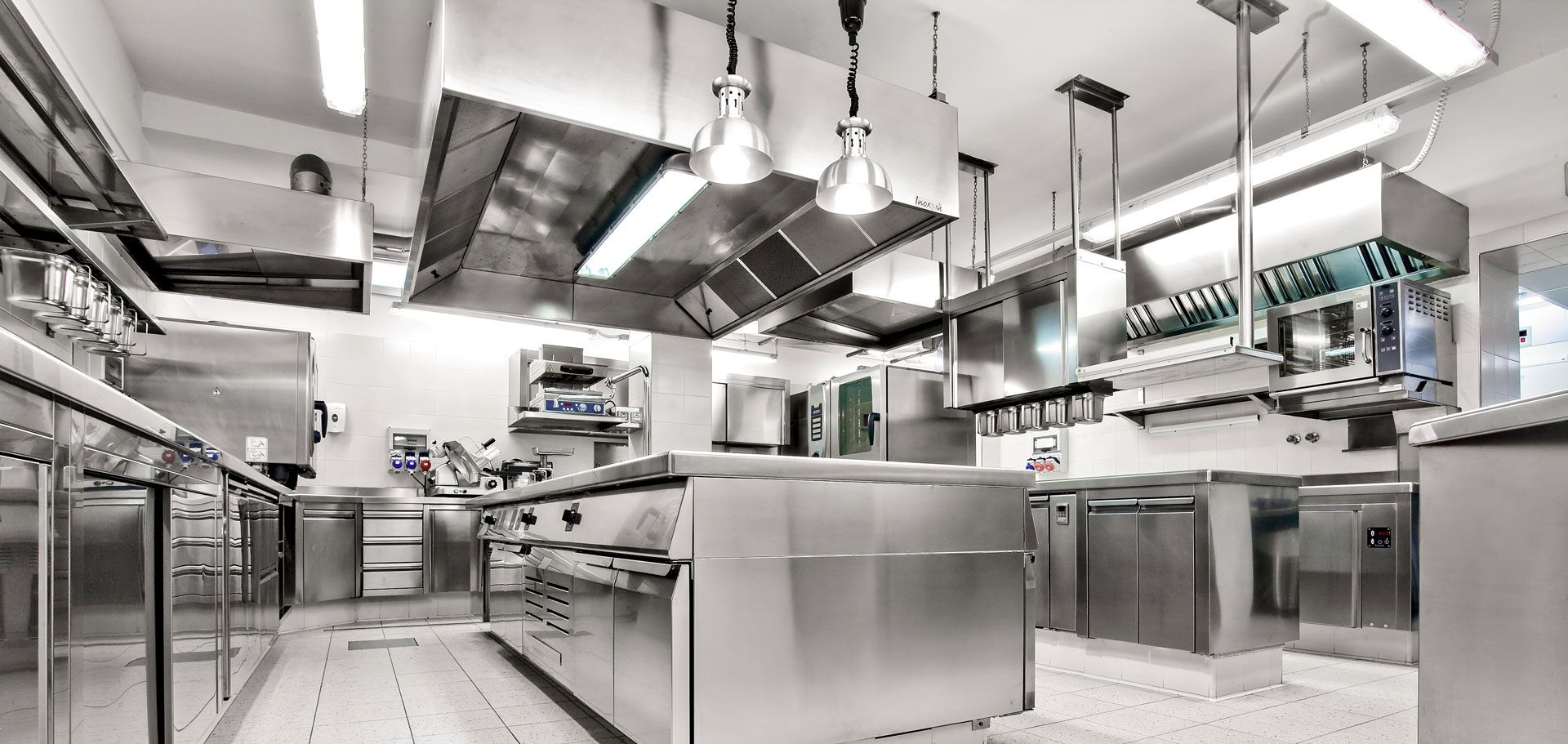 Modern stainless-steel commercial kitchen with counters, ovens, and hanging pots under bright lights