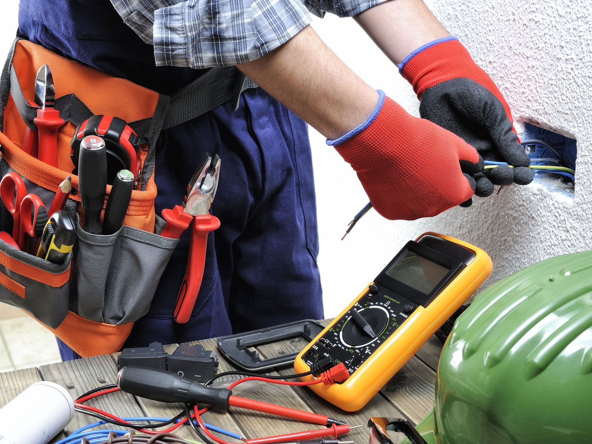 Electrician working on electrical outlet, wearing work gloves and tool belt, with multimeter and helmet on a table.