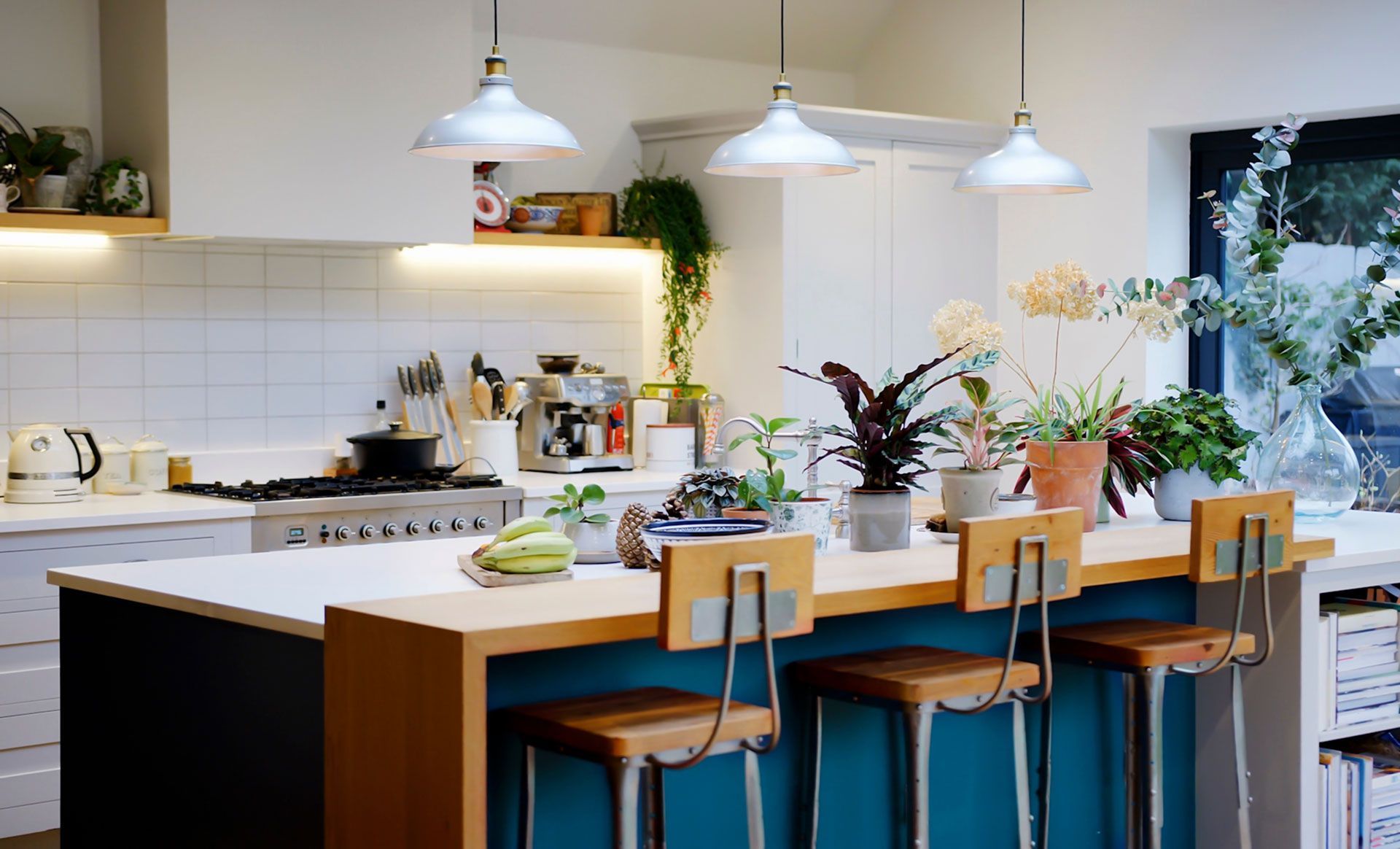 Modern kitchen with a wooden island, blue stools, hanging lights, and plants by a bright window