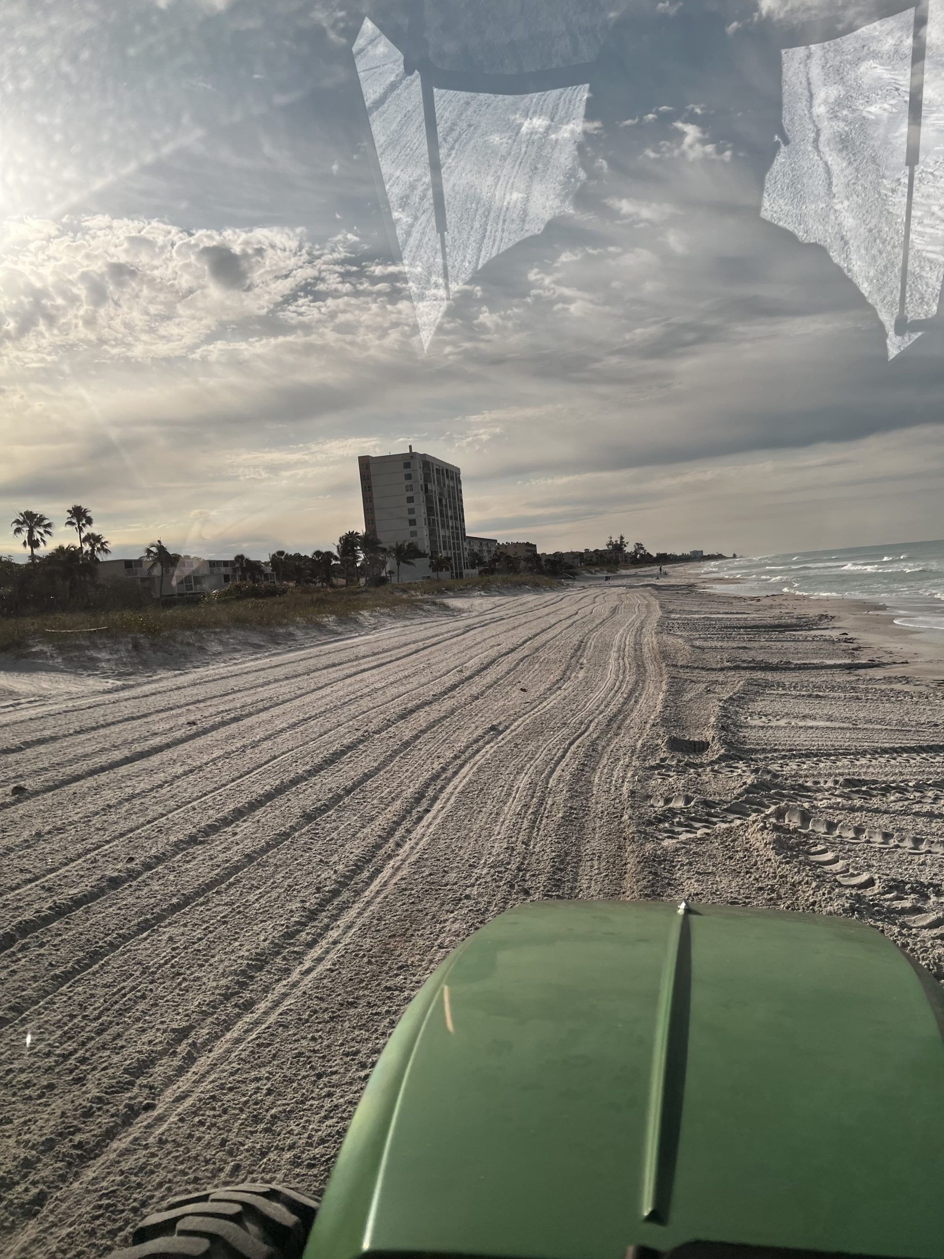View from a green tractor driving on a beach, with buildings and ocean visible in the distance under a cloudy sky.