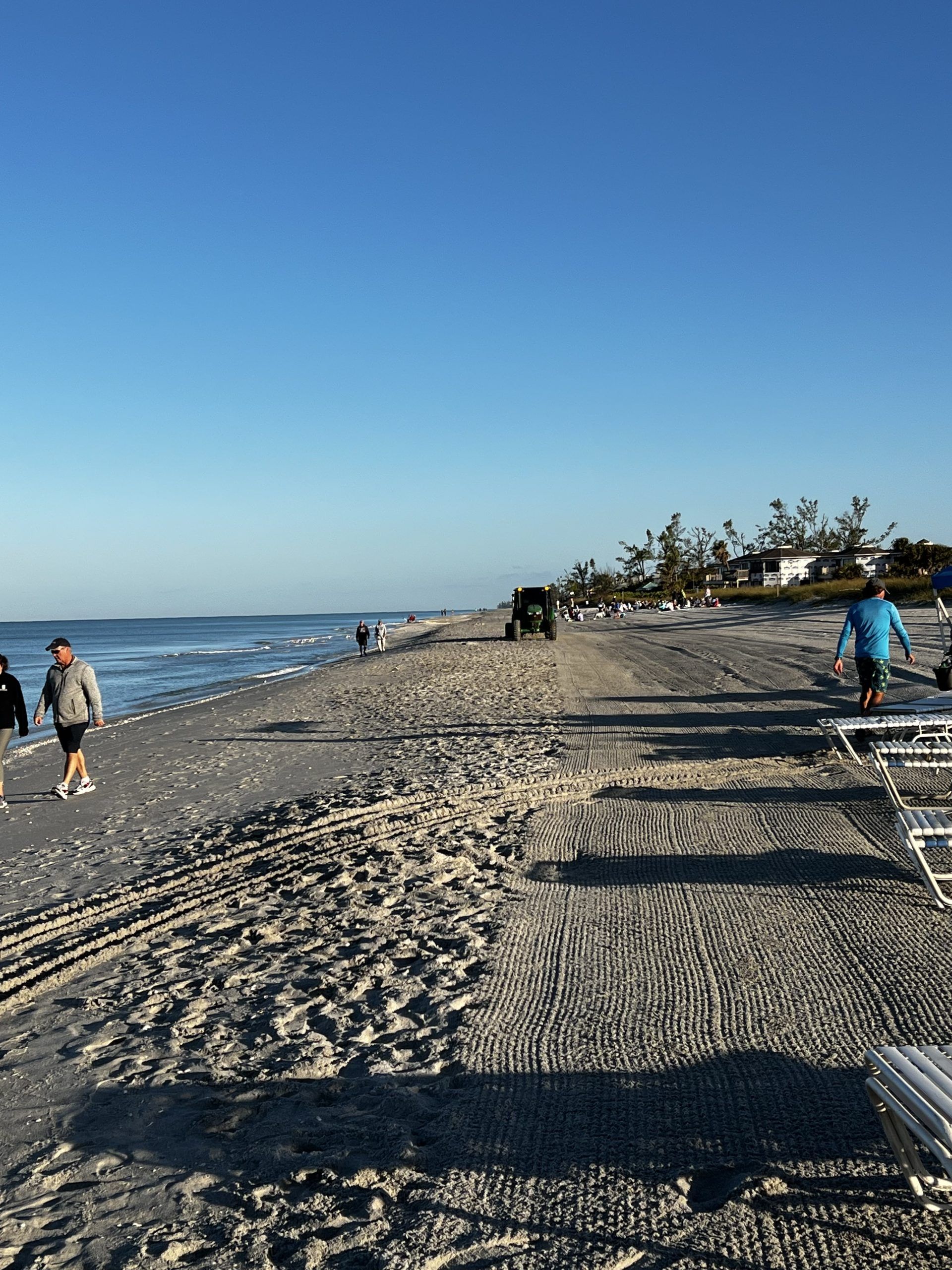Beach scene with people walking on sand; blue sky and water.