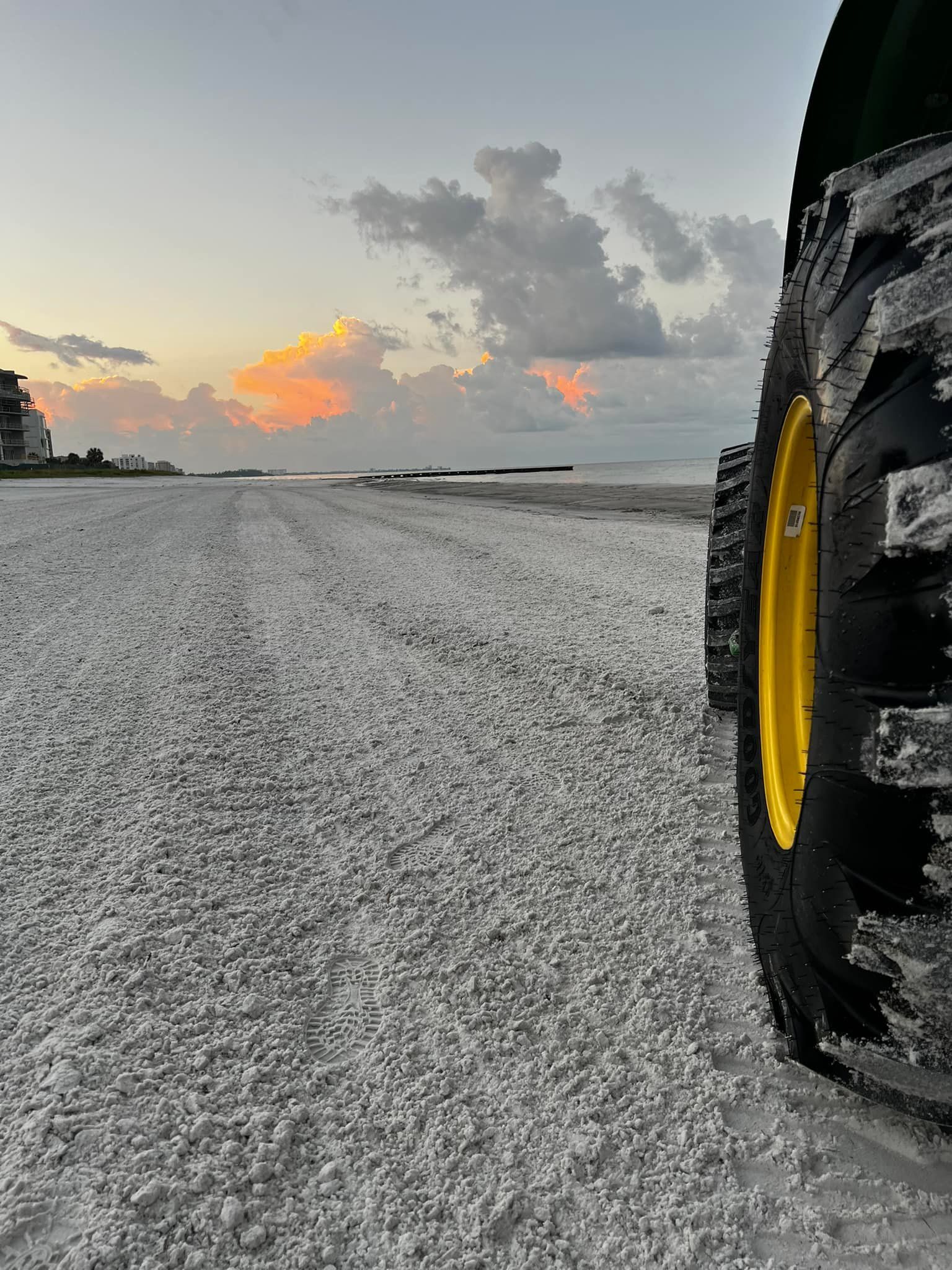 Tractor tire on a shell-covered beach at sunset, with buildings in the distance and cloudy sky.