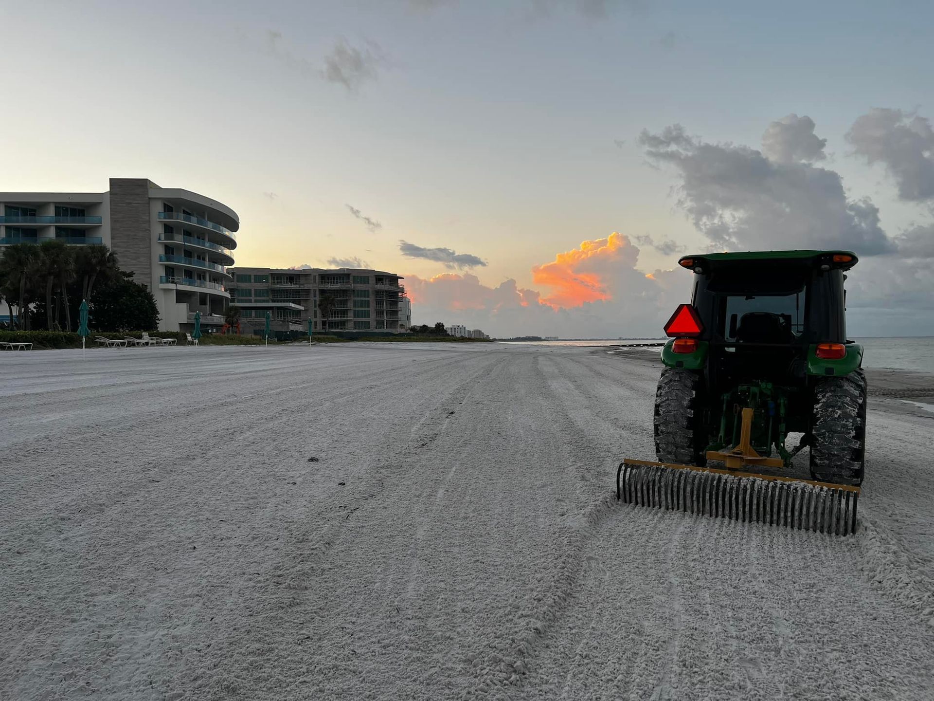 A tractor rakes a sandy beach at sunrise, with a hotel in the background.