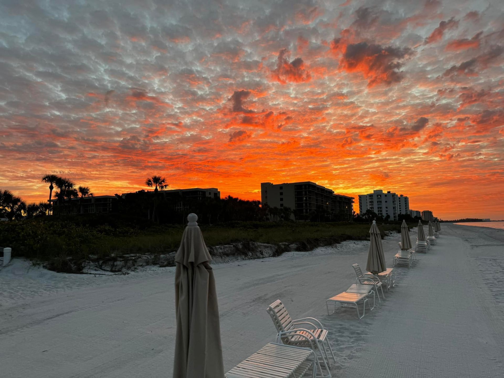 Beach at sunset with vibrant orange sky, lounge chairs, and buildings silhouetted in the distance.
