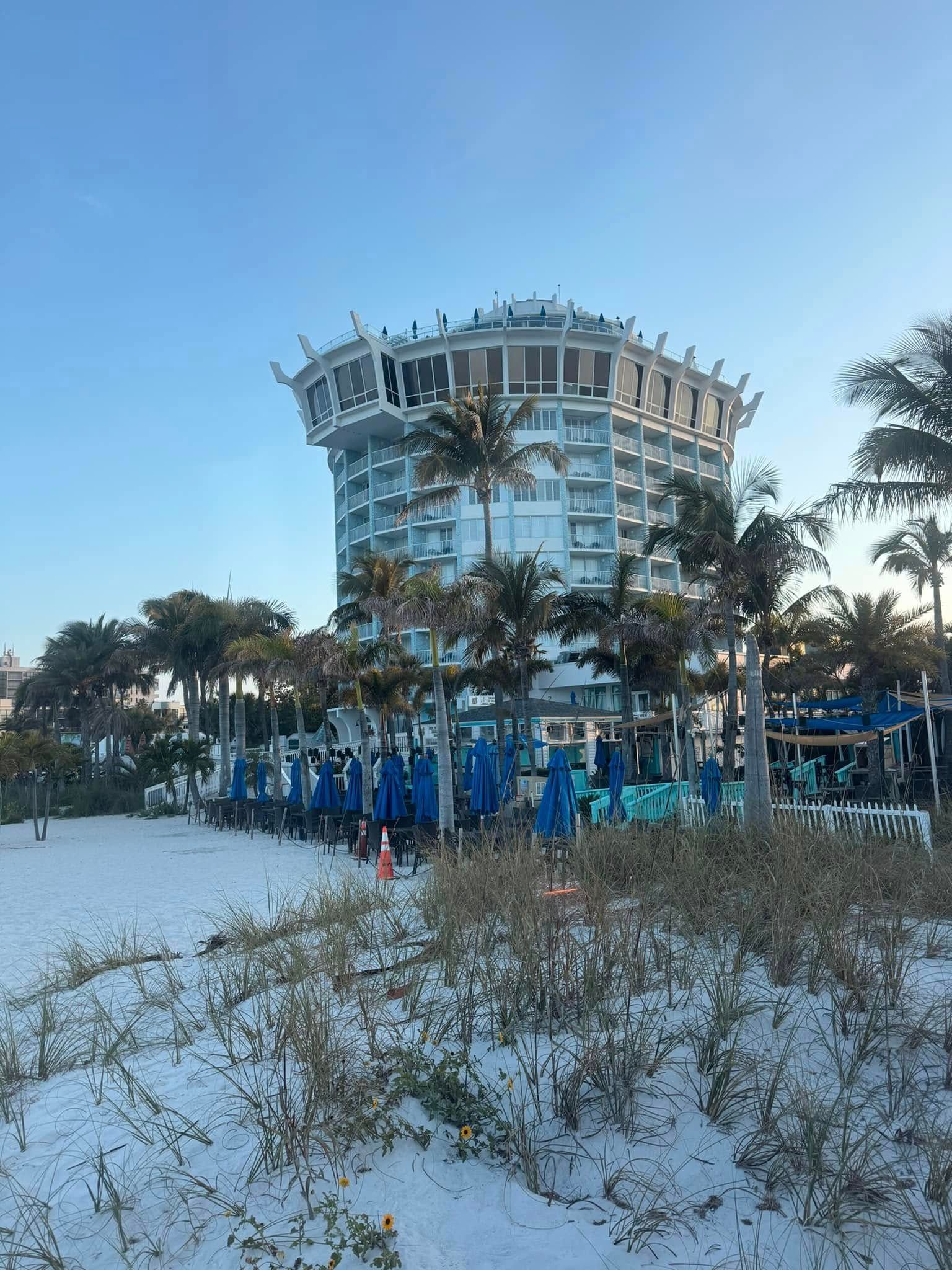 Beach view with a round, white hotel surrounded by palm trees and blue umbrellas.