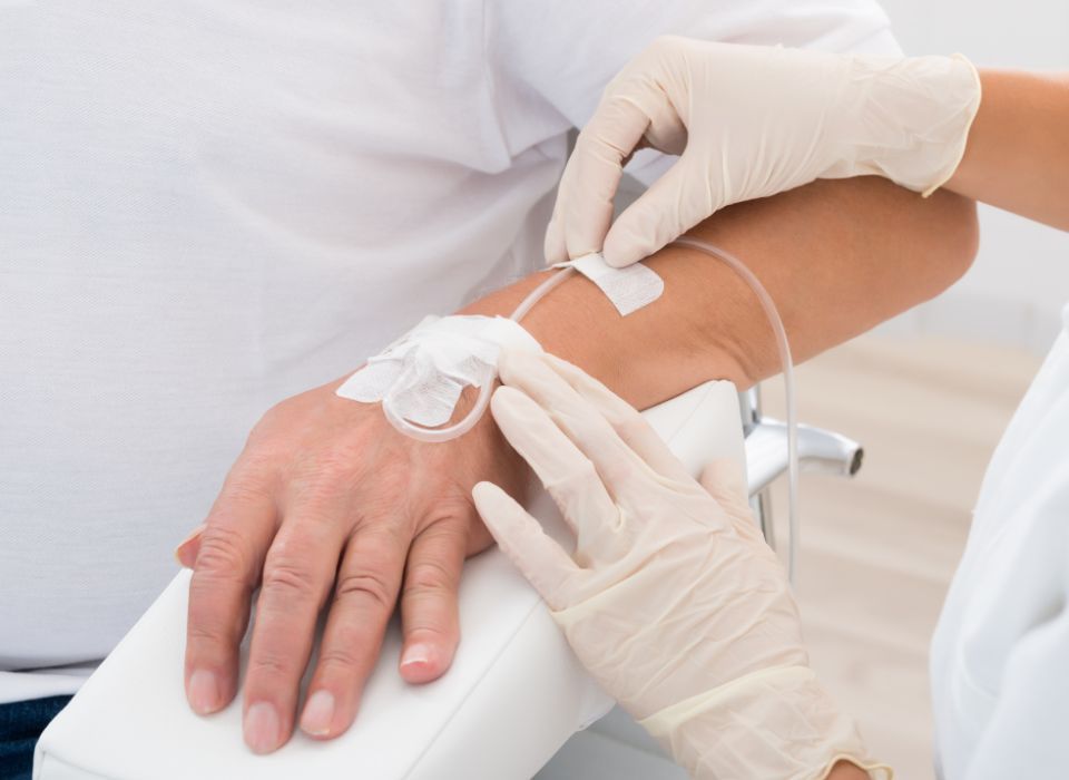 A person receiving an IV drip in their arm, held by a healthcare worker wearing gloves.