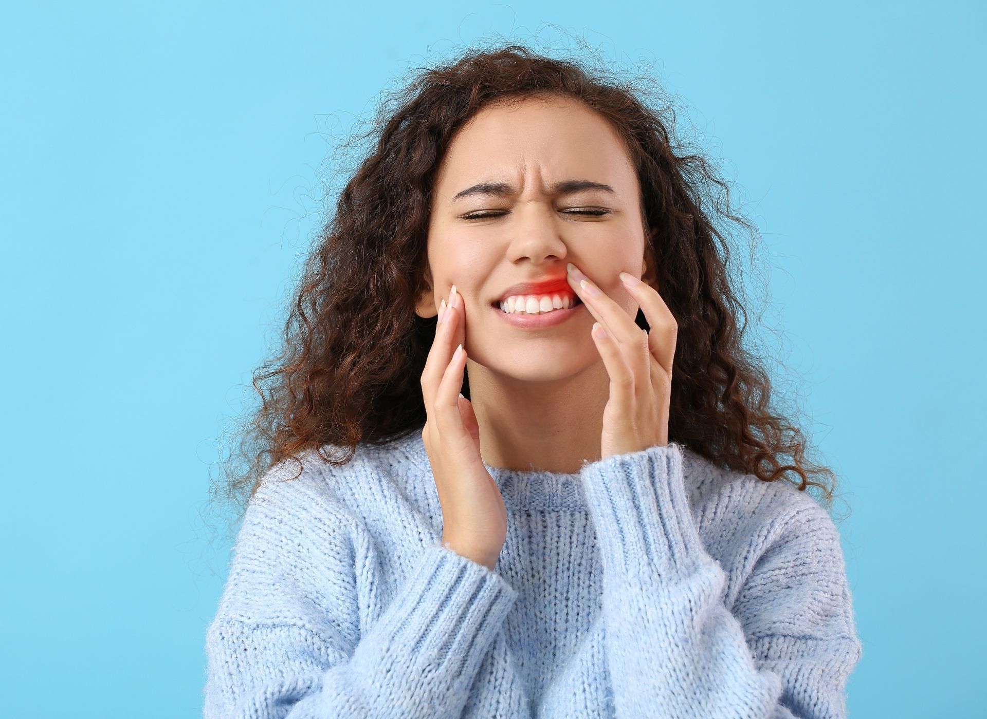 Woman with curly hair grimaces, touching her cheek and lip, displaying red, irritated gums.