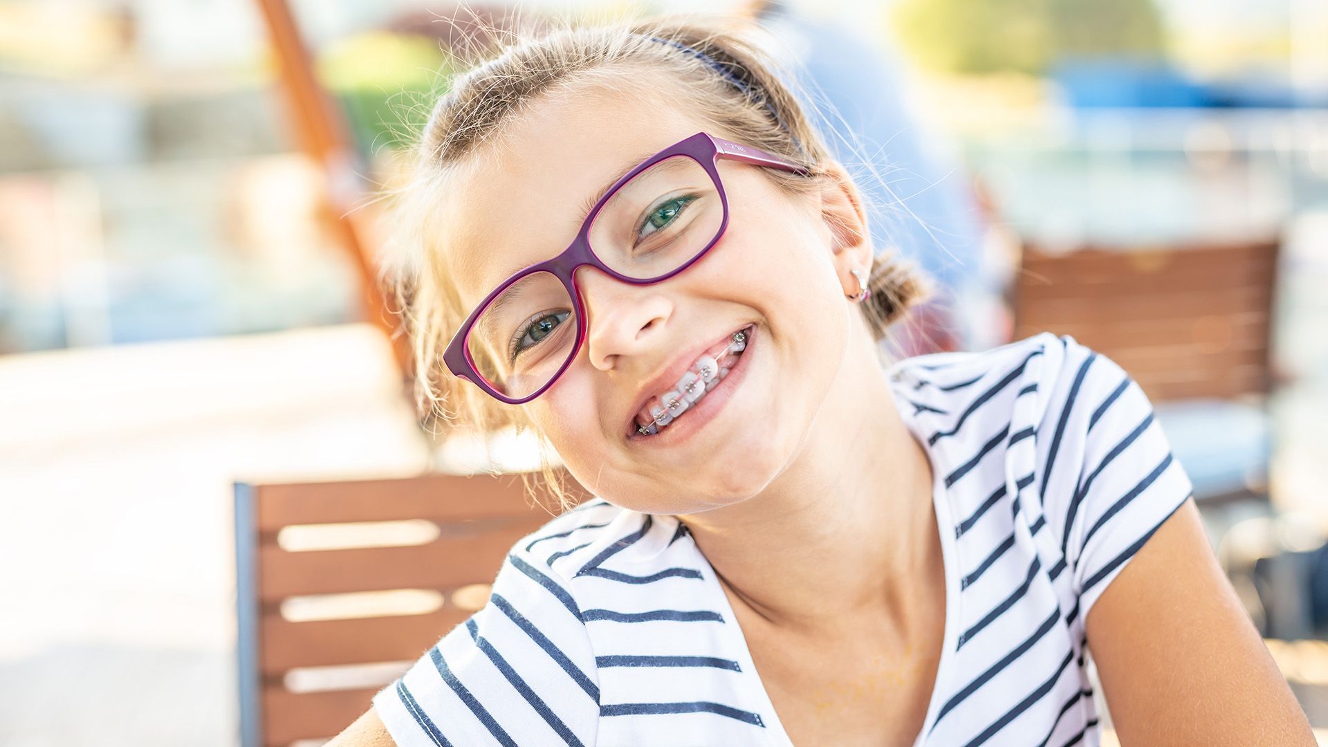 Smiling person wearing purple glasses and striped shirt, leaning outdoors in bright sunlight.