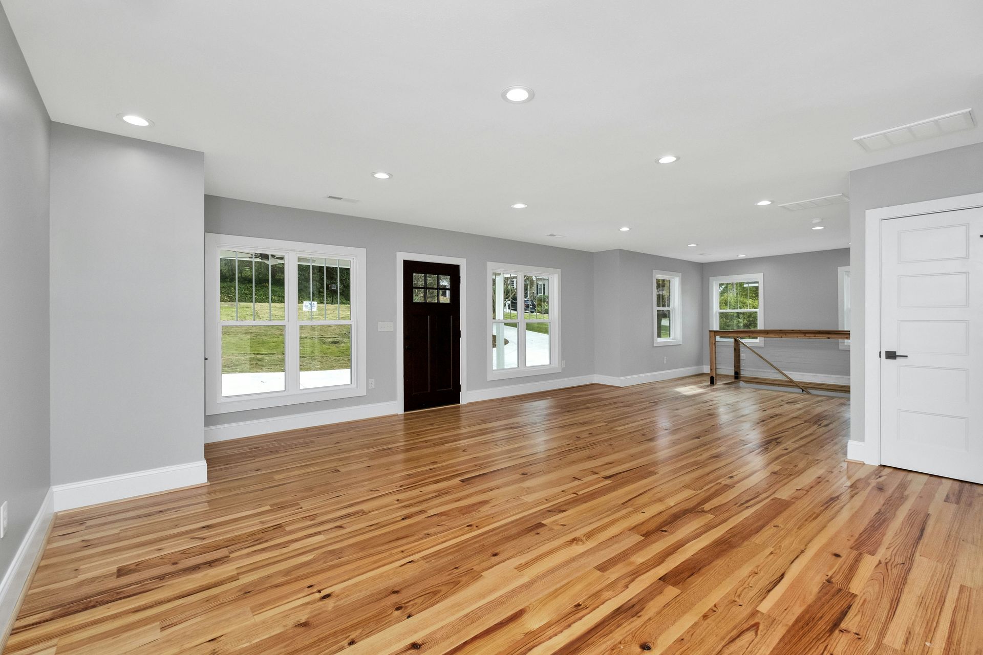 Empty modern living room with hardwood floors, gray walls, large windows, and recessed ceiling lights