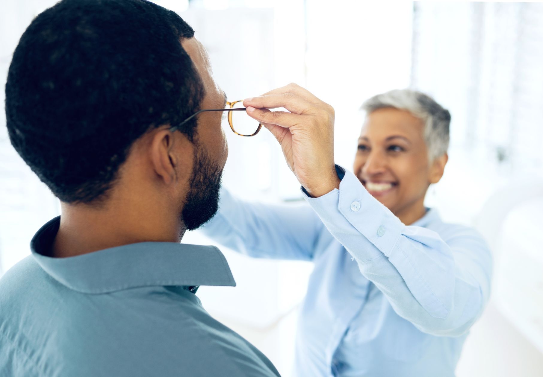 A woman is helping a man try on glasses in front of a mirror.