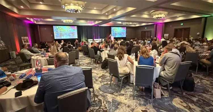Conference banquet hall with seated attendees, round tables, and presentation screens at the front