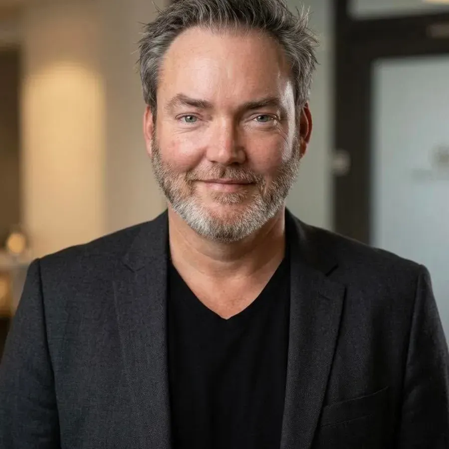 Man smiling in a dark blazer and black shirt, standing indoors with a softly blurred background