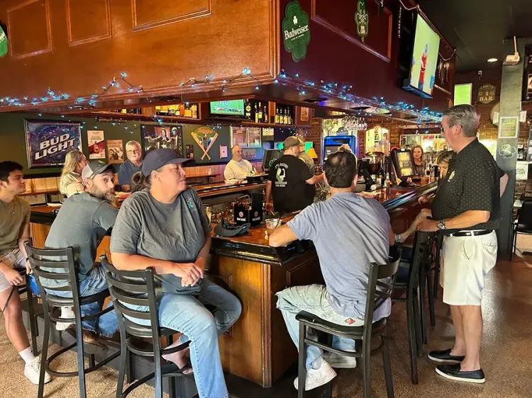 Patrons sit at a busy bar with TV screens and neon signs in a dimly lit pub.