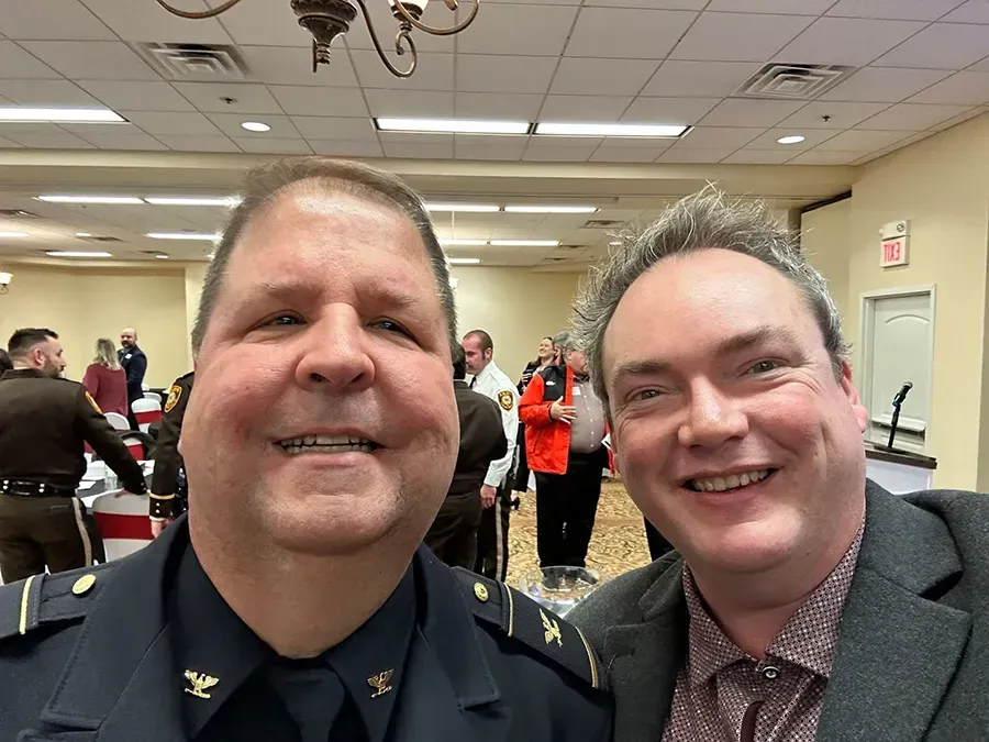 Two smiling men posing for a selfie at an indoor event with people and tables in the background