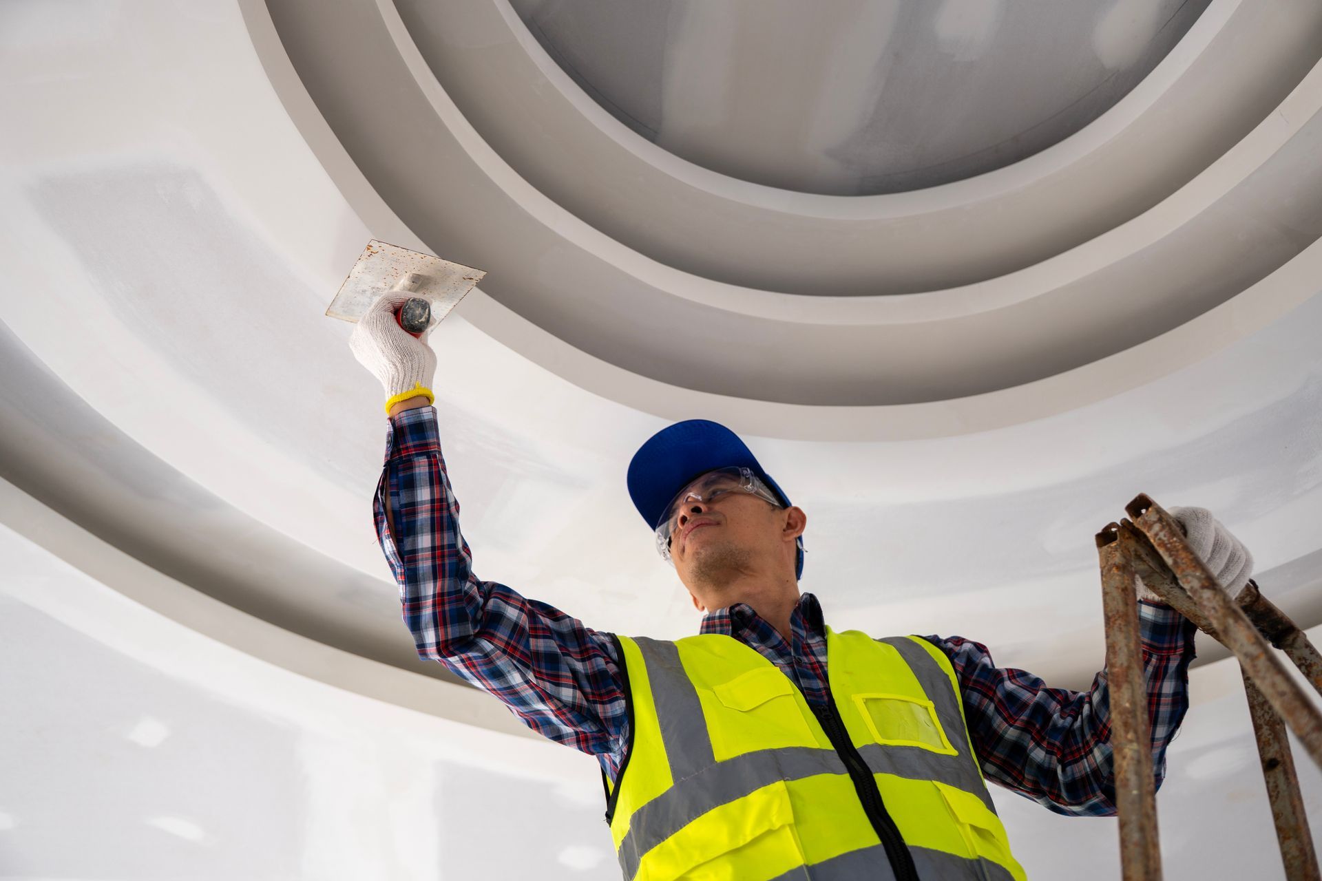 Worker in a safety vest paints a white ceiling while standing on a ladder.