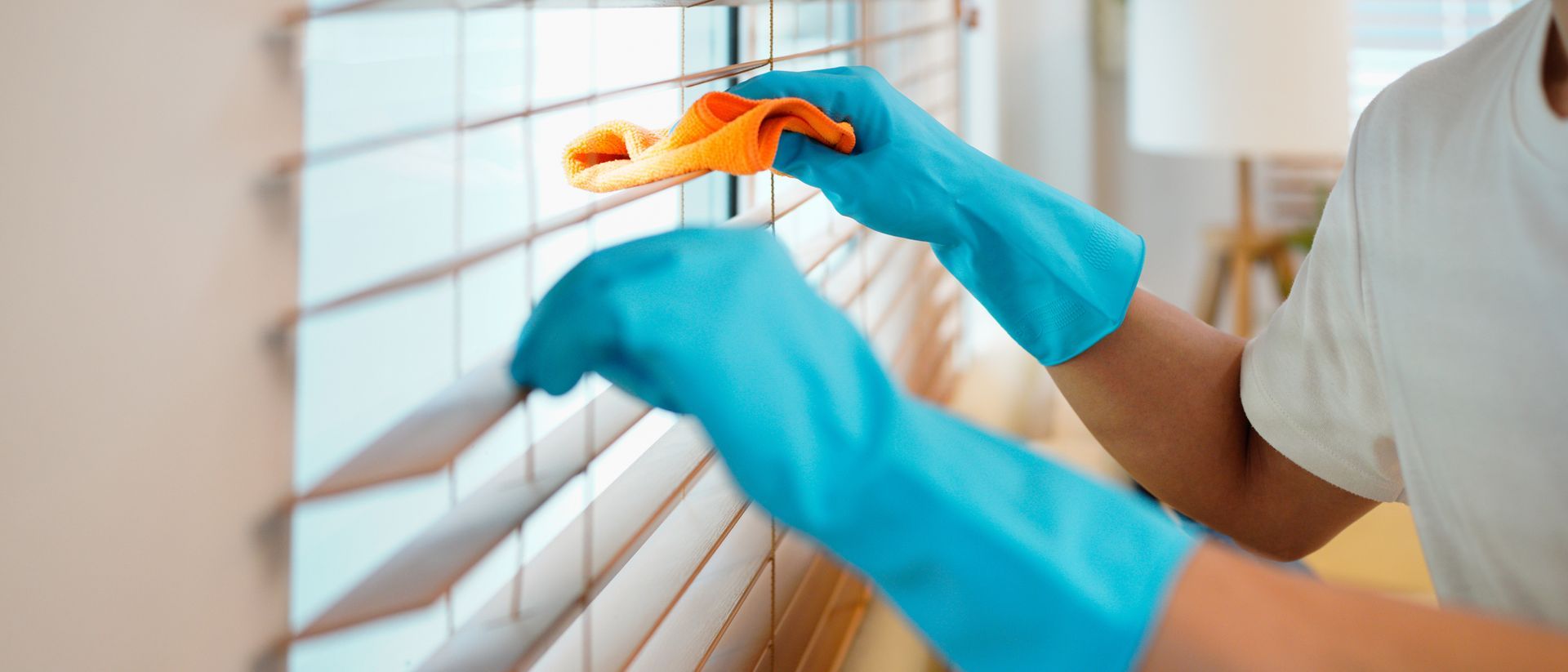 Blue-gloved hands cleaning window blinds with an orange cloth in a bright room