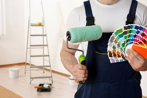 Person holding paint roller and color swatches in a room with a ladder and paint supplies