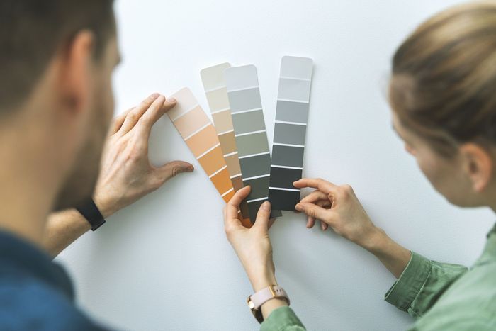 Two people compare paint swatches against a white wall, choosing neutral and warm color samples.