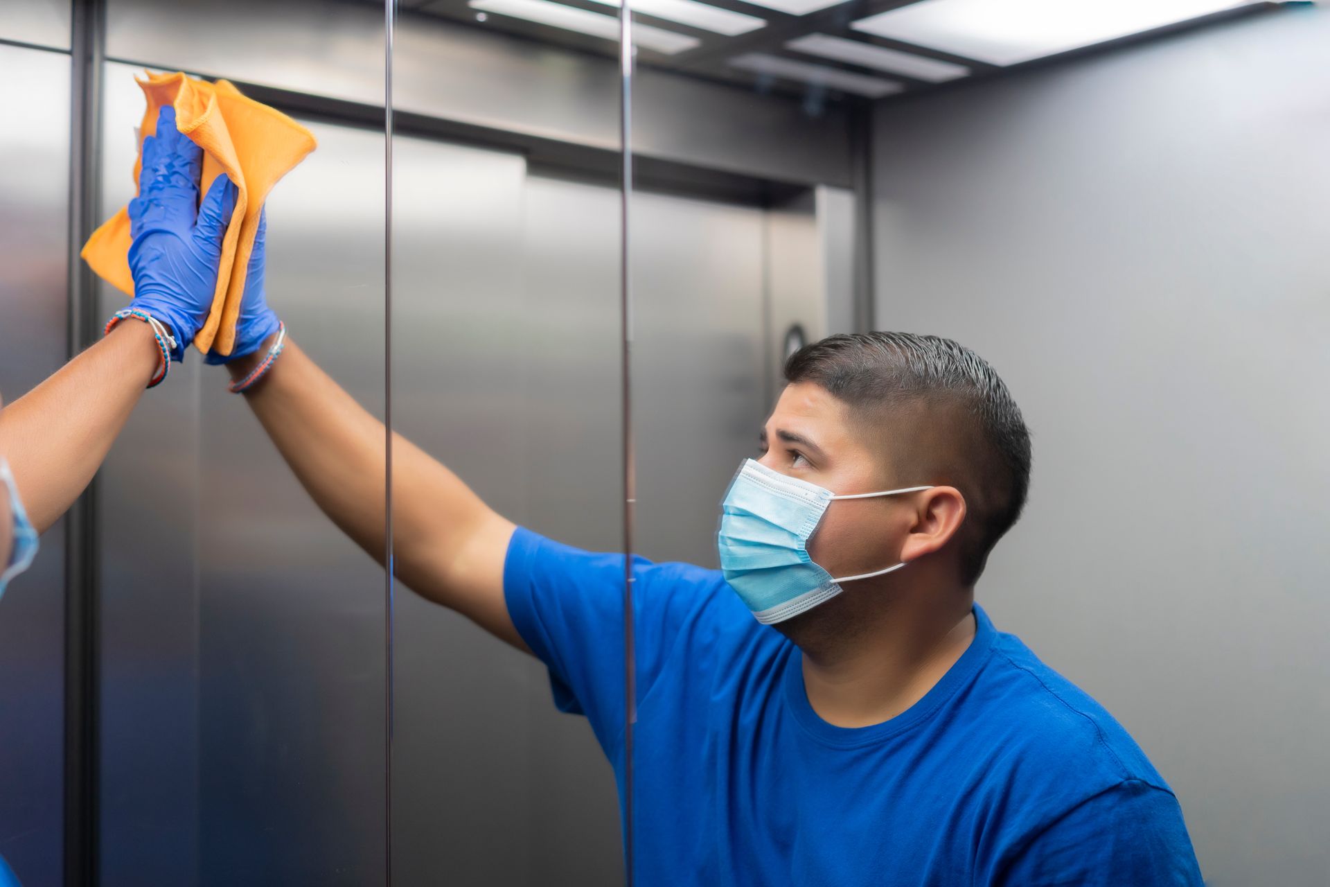 Masked cleaner wiping an elevator wall with a cloth while standing inside the elevator