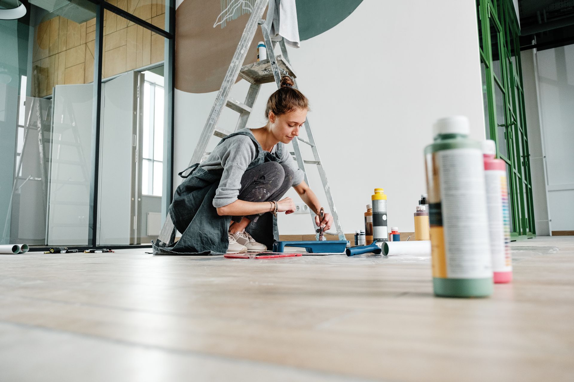 Person crouching on a wooden floor, painting or repairing a wall in a bright room with tools and ladders nearby Person crouching on a wooden floor, painting or repairing a wall in a bright room with tools and ladders nearby