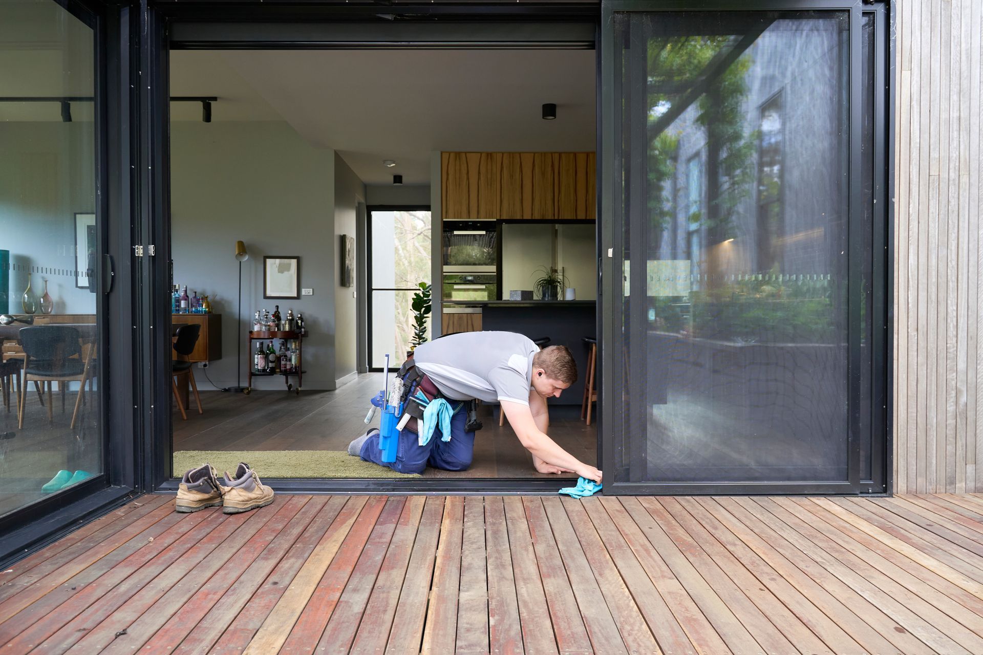 Person kneeling on a wooden deck cleaning a sliding glass door beside a modern patio. Person kneeling on a wooden deck cleaning a sliding glass door beside a modern patio.