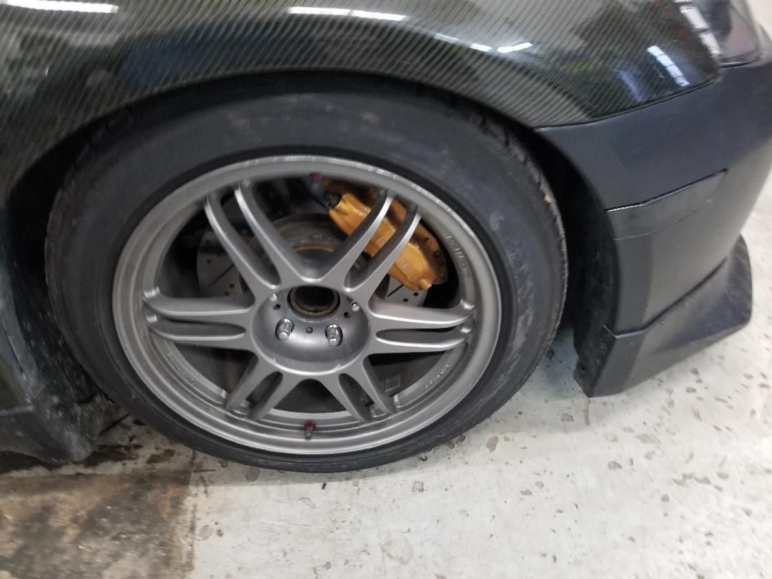 Close-up of a black car’s front wheel with gray alloy rim and visible orange brake caliper on a concrete floor.
