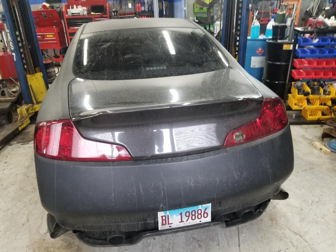 Rear view of a gray sedan in an auto shop with a damaged bumper and visible license plate.