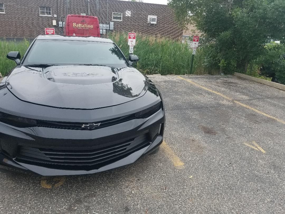 Black sedan parked in a gravel lot with trees and a red truck in the background