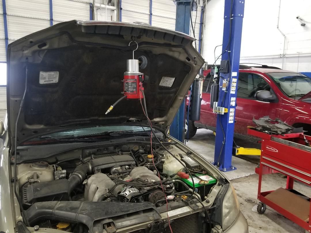 Car engine bay open in an auto shop, with a hood prop light and a blue lift nearby.