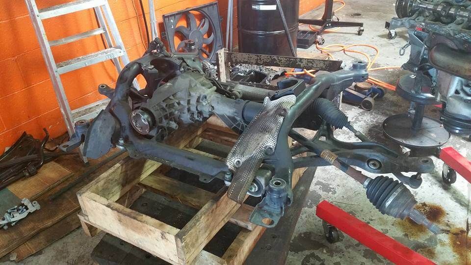Rusty vehicle axle assembly on a wooden pallet in a workshop, with tools and metal parts nearby