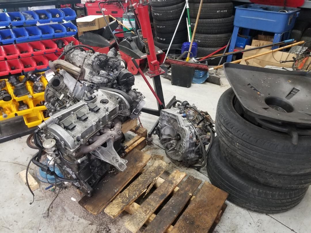 Two dirty engine parts on a pallet in a cluttered workshop beside stacked tires.