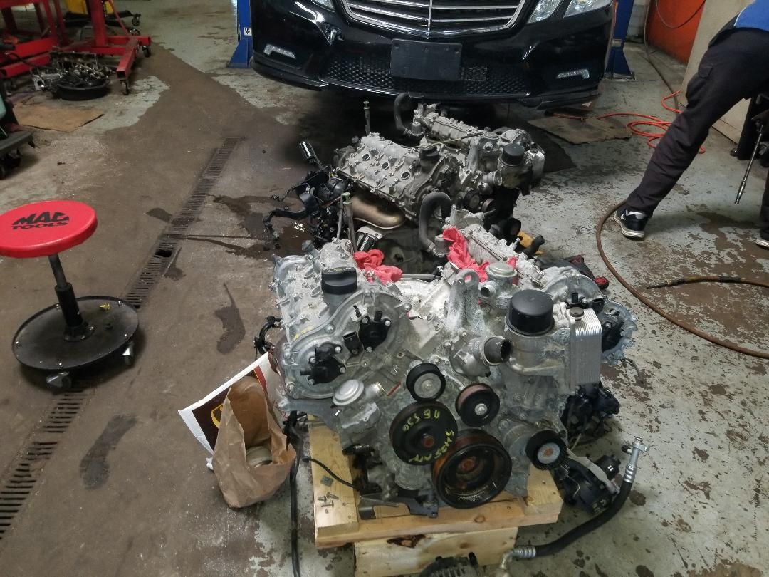 Car engine parts arranged on a pallet in a garage, with a black car in the background.