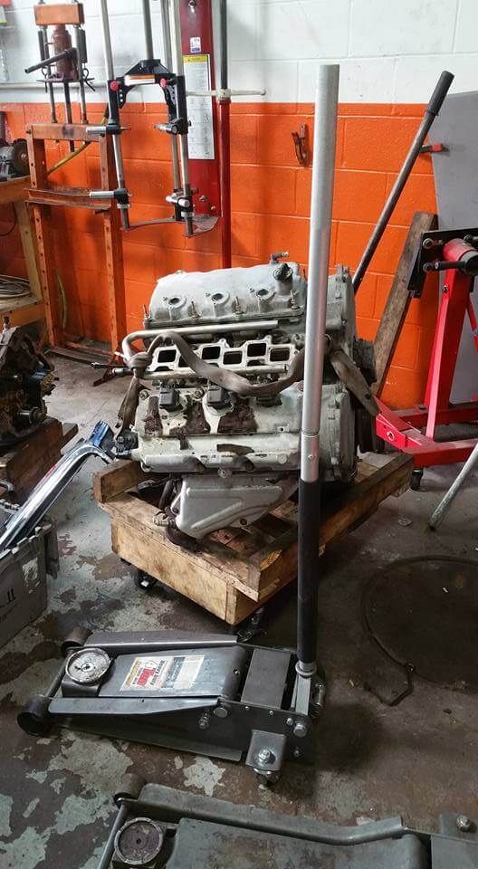 Engine block on a floor jack in a cluttered auto shop with orange walls and tools nearby