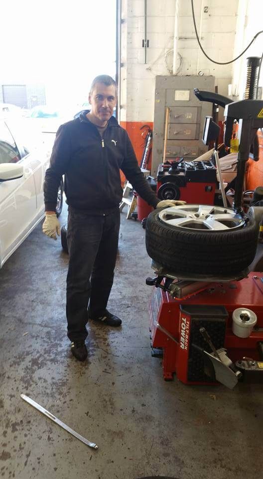 Mechanic standing beside a tire machine in an auto repair shop, with tools and vehicle parts around him