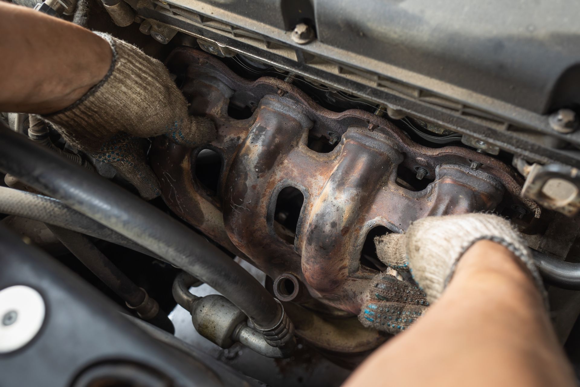 Gloved hands removing a rusty car exhaust manifold in an engine bay