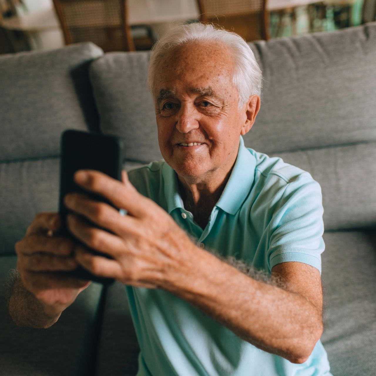 An elderly man is sitting on a couch taking a selfie with his cell phone.